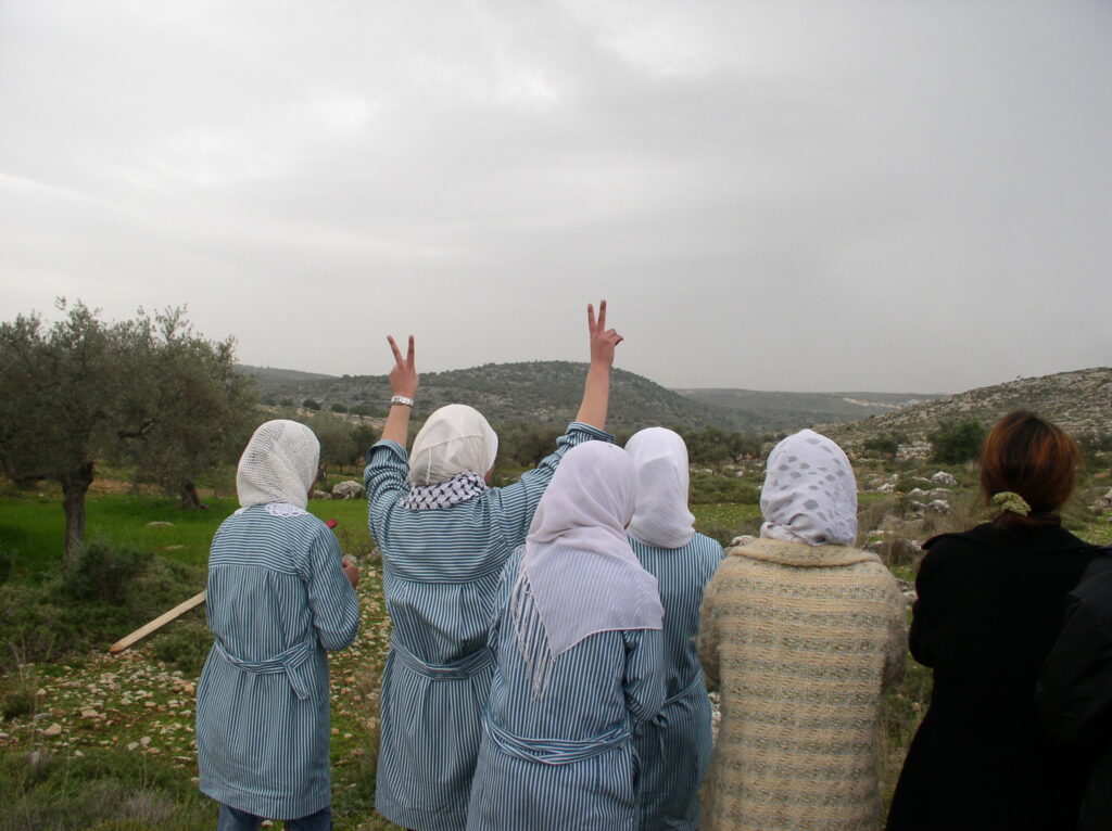 A photo of four female students and two female adults with their backs to the camera. Student in the middle is holding up peace signs with both her hands.