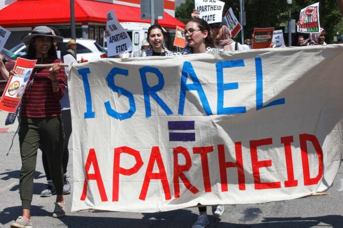 Photo of protesters marching in the street holding a banner that reads "Israel = Apartheid" with Israel in light blue, = in purple, and apartheid in red, all caps, on a white background. The protested appear to be women of varying ethnicities, some wearing keffiyahs. Protesters hold other posters, too, including "Israeli is an apartheid state," "freedom for Gaza" with a rising fist," and "boycott, divest, sanction."